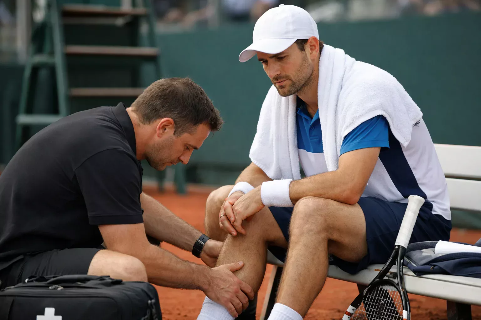 Tennista seduto a bordo campo con asciugamano mentre il fisioterapista valuta un infortunio alla gamba